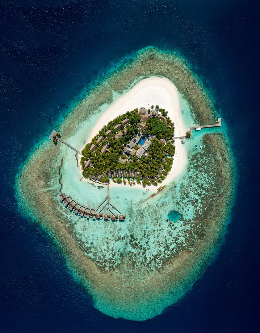 Kandolhu Maldives, aerial, Alif Alif Atoll (North Ari), Maldives, Top-down drone aerial of the boutique triangular Kandolhu island, the 30-villa thatched cluster spread across the palm-canopied small island with the twin jetties projecting at one end and the reef-edge halo encircling. The boutique-island intimate scale signature.