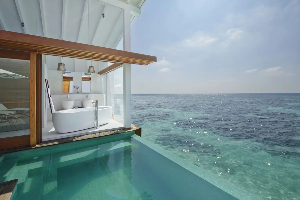 Kandolhu Maldives, villa overwater, Alif Alif Atoll (North Ari), Maldives, Ocean Pool Villa interior, open-side bathroom with the freestanding tub on a marble platform overlooking the infinity-edge cantilevered plunge pool that runs the deck edge, bright marine light flooding through the open-side villa configuration.