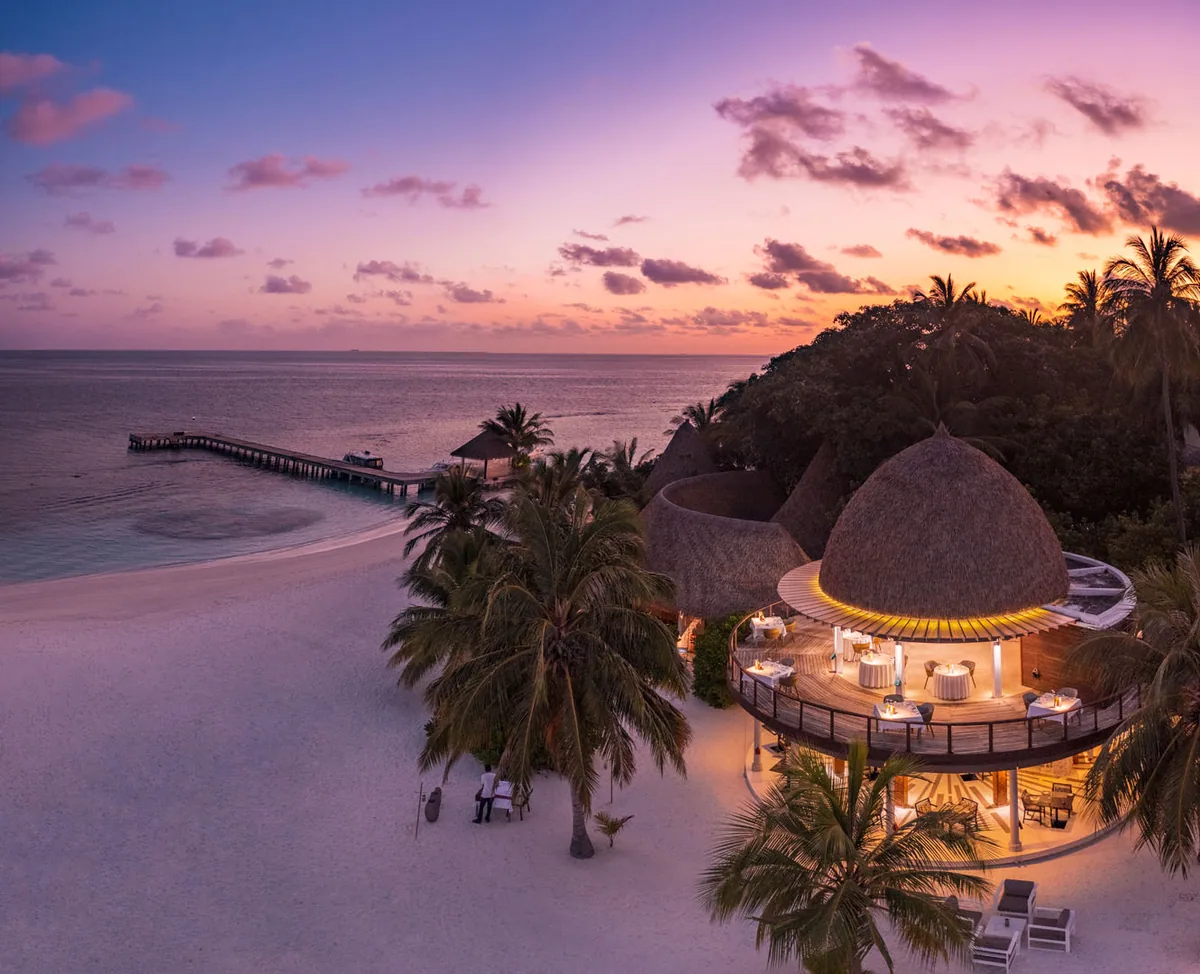 Kandolhu Maldives, dining, Alif Alif Atoll (North Ari), Maldives, The Market and Olive at dusk, double-tier circular thatched-roof open-air dining pavilion lit warmly from within, long timber jetty extending into the magenta-amber sky reflection on the lagoon. The property's principal venue in cinematic composition.