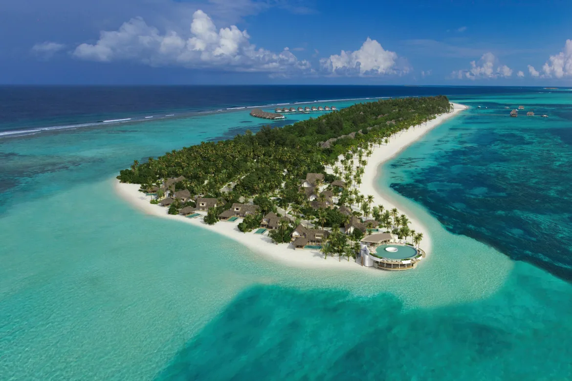 Kanuhura Maldives, aerial, Lhaviyani Atoll, Maldives, Six Senses Kanuhura drone aerial: the long narrow one-kilometre Kanuhura island stretching toward the horizon, white sand beach perimeter, beach pool villas clustered along the spine of the natural-vegetation island, signature spa pod with circular plunge pool at the foreground, OWV cluster at the far end of the island, encircling turquoise lagoon, Lhaviyani Atoll.