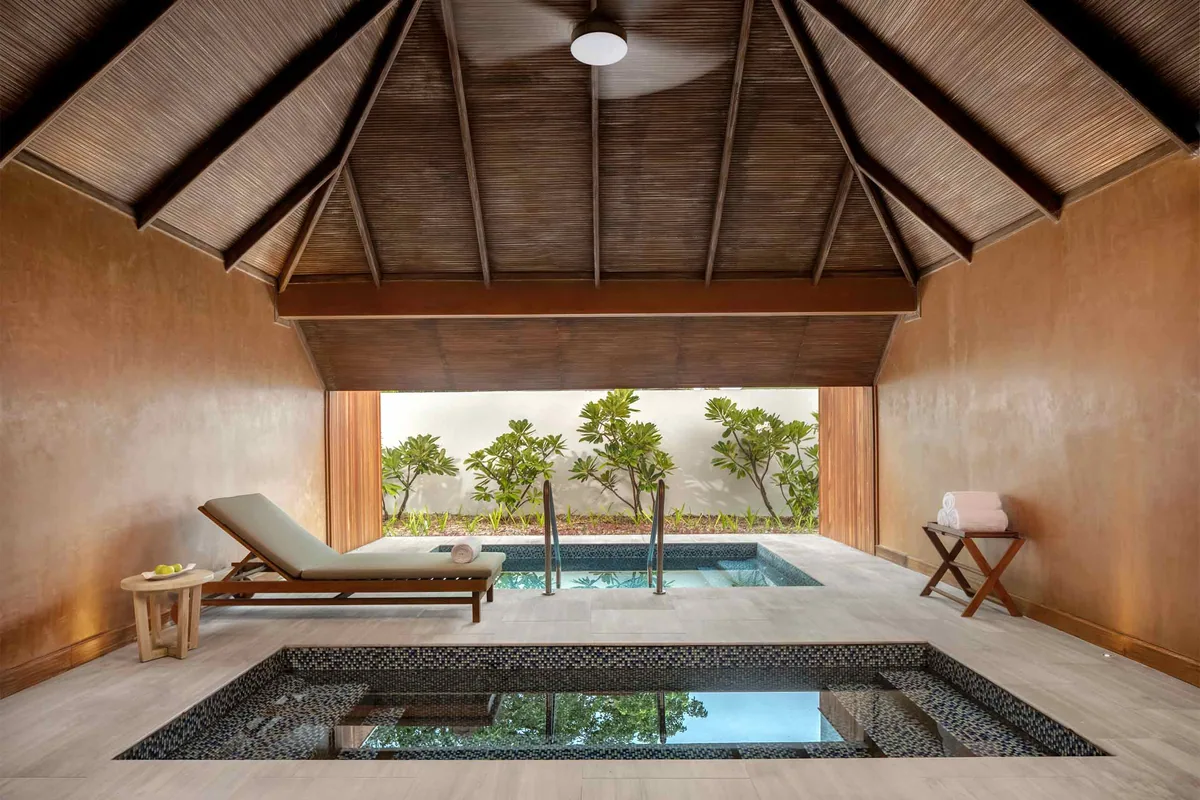 Kanuhura Maldives, spa, Lhaviyani Atoll, Maldives, Six Senses Kanuhura spa treatment pavilion: vaulted timber rafter ceiling, central glass-tile plunge pool with mosaic floor, sun lounger at left, terracotta-clay walls, open frontage onto a small plumeria-tree garden courtyard.