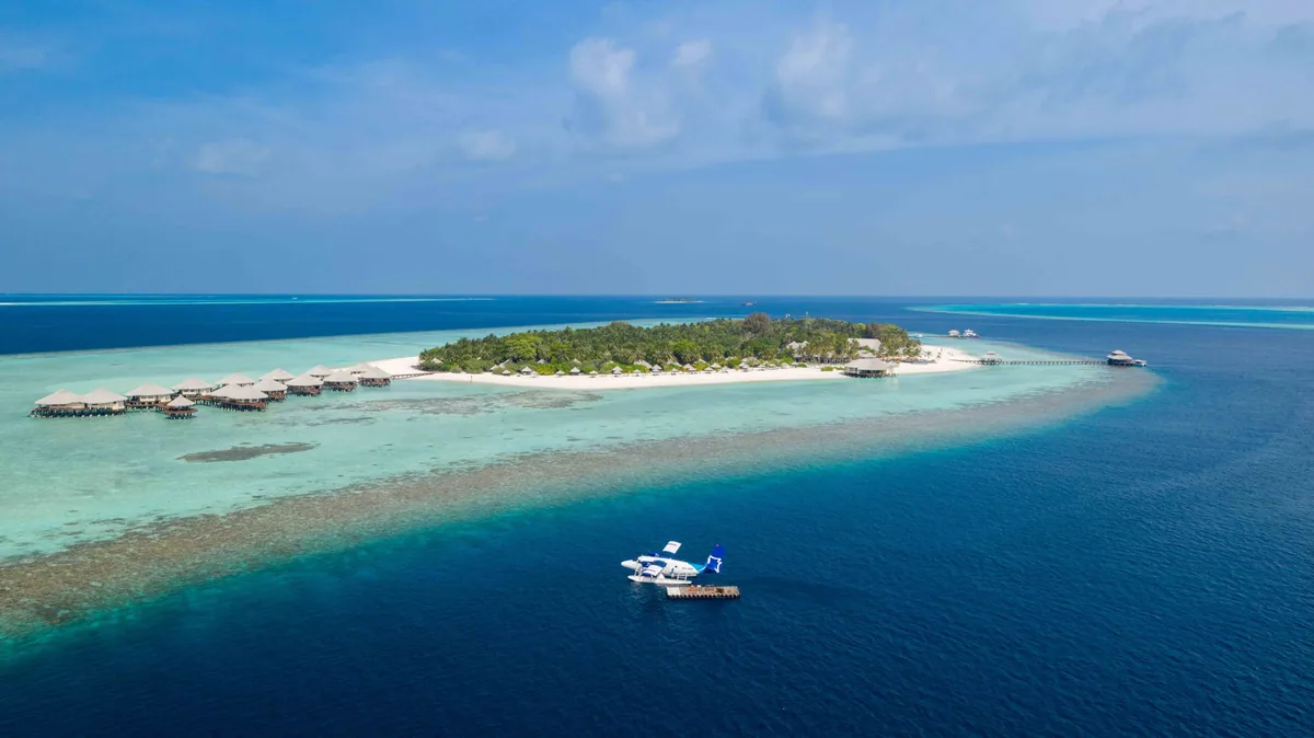 Kihaa Maldives, aerial, Baa Atoll, Maldives, Kihaa Maldives from the drone height, the long coconut-shaped Kihaadhuffaru island geometry with the fern-pattern overwater villa cluster reaching off the western lagoon arm, the Trans Maldivian seaplane docked at the foreground jetty and the secondary arrival jetty extending out the eastern reef shelf.