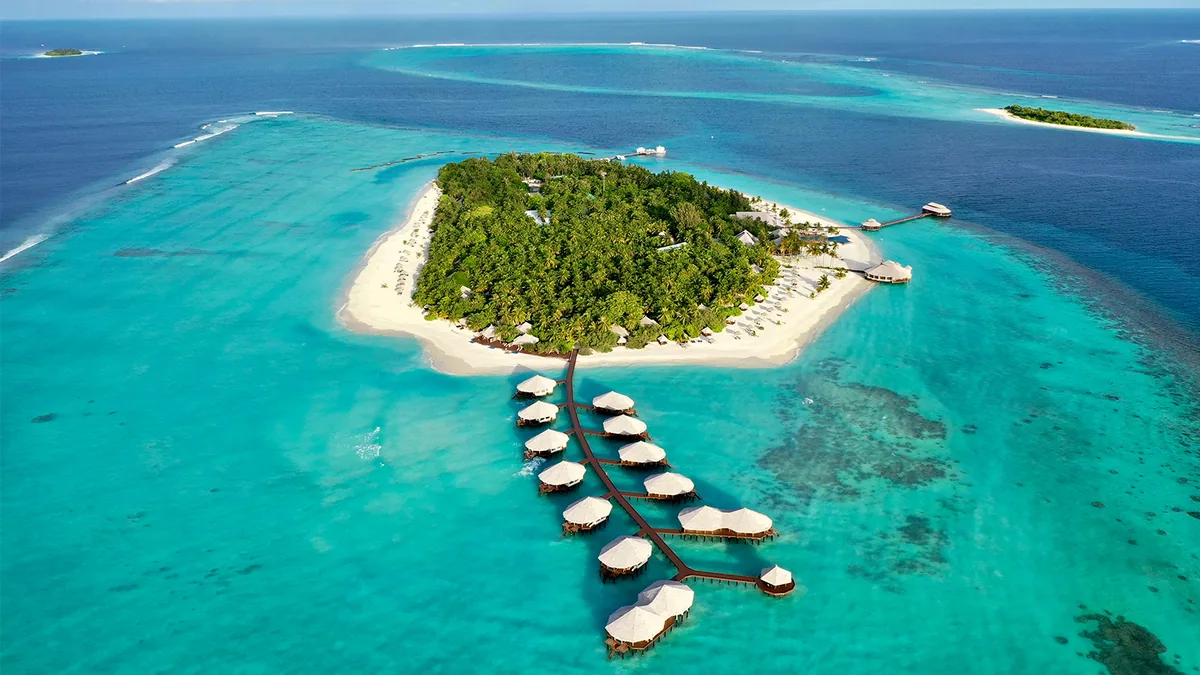 Kihaa Maldives, setting, Baa Atoll, Maldives, Fern-pattern overwater villa cluster reaching off the eastern jetty arm, the rows of thatched-conical roof villas branching from the central boardwalk like a fern skeleton, the surrounding turquoise reef shelf and the outer-atoll dark blue water with concentric reef rings beyond.