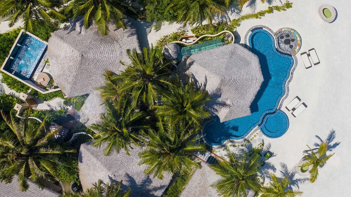 Kihaa Maldives, villa beach, Baa Atoll, Maldives, Royal Suite top-down drone view, the curved kidney-shape mosaic-tile private pool with twin cushioned daybeds at the apex, the paired thatched-conical villa pavilions with the deck connecting to the white-sand beach edge under dense coconut-palm canopy.
