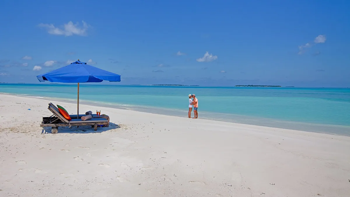 Kihaa Maldives, activities, Baa Atoll, Maldives, Private sandbank excursion, a couple in casual sun-clothes embracing at the water's edge of a sun-bleached sandbank with the signature Kihaa blue parasol and the wooden sun-lounger set with welcome drinks, the long line of distant Baa Atoll islands strung across the horizon.
