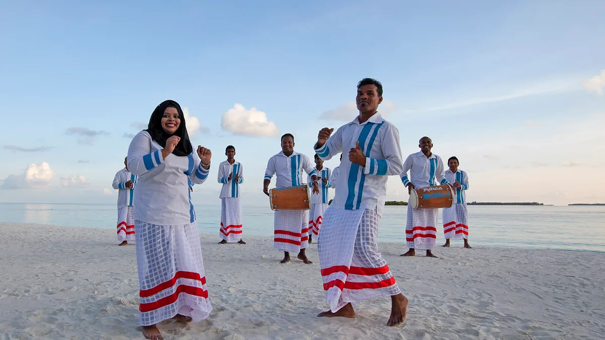 Kihaa Maldives, activities, Baa Atoll, Maldives, Bodu Beru traditional drum-and-dance performance on the beach at the southwest-monsoon dusk, the Maldivian dance troupe in white shirts with cobalt-blue chest-stripes and red-banded white-check sarongs, three Kihaa-branded barrel drums and the calm lagoon stretching to the horizon behind.