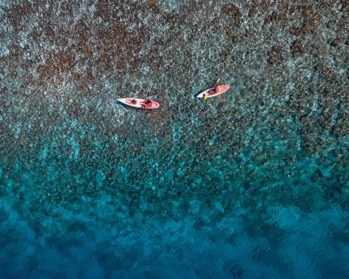 Kihaa Maldives, activities, Baa Atoll, Maldives, Two single kayaks drifting over the Kihaadhuffaru house-reef from the drone-height view, the gradient blue-to-deep-blue of the reef shelf 200 metres off the Hanifaru Bay MPA boundary, the underwater coral pattern visible in clean detail under the sun-dappled surface.