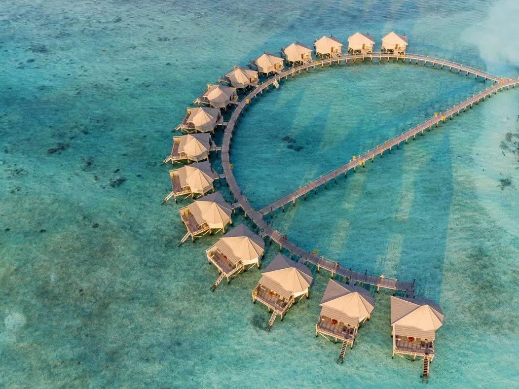 Komandoo Island Resort, villa overwater, Lhaviyani Atoll, Maldives, Water Villa cluster full top-down aerial: the distinctive U-shape arc of approximately sixteen thatched-pyramid-roof overwater villas suspended on stilts along a curving timber walkway, the crystal-turquoise lagoon and reef pattern visible beneath, the signature Komandoo overwater-villa-arc geometry.