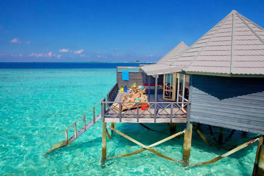 Komandoo Island Resort, villa overwater, Lhaviyani Atoll, Maldives, Individual Water Villa ground-level perspective: signature thatched-pyramid roof with rolled-edge profile, timber-clad walls, X-bracing structural stilts in the crystal lagoon, the private deck with twin sun loungers and the step-down ladder descending directly into the lagoon, the distant atoll horizon framing the composition.