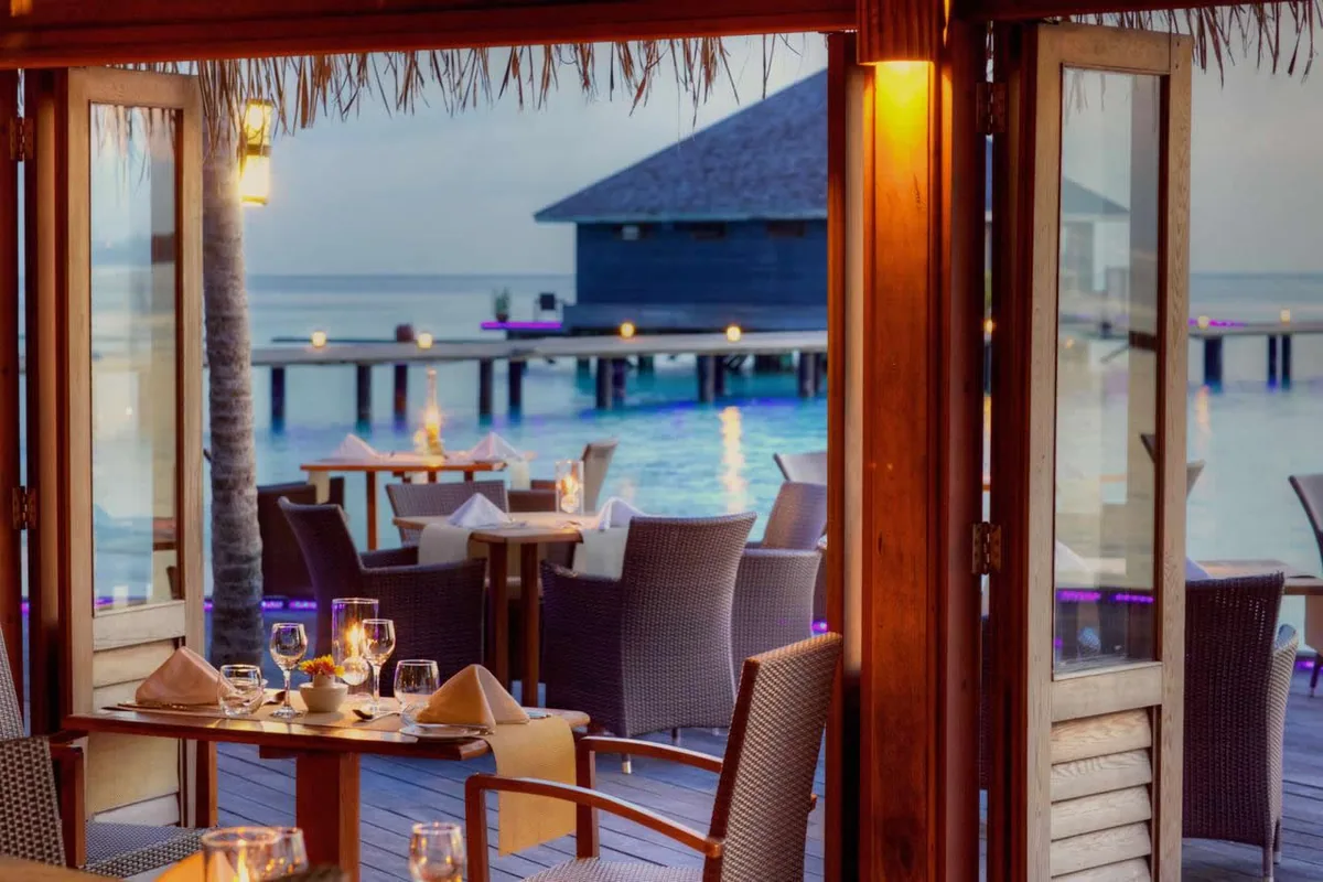 Komandoo Island Resort, dining, Lhaviyani Atoll, Maldives, Aqua overwater dining pavilion at twilight: view through wooden bi-fold glass doors framing the candle-lit tables with dark wicker chairs, palm trunk integrated through the interior, the calm lagoon water and the overwater pavilion plus jetty visible beyond, warm amber lantern lighting against the cool dusk sky.