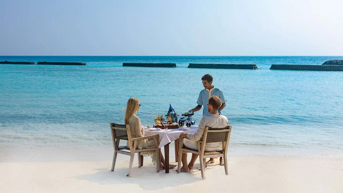 Komandoo Island Resort, dining, Lhaviyani Atoll, Maldives, Beachside private dining at the lagoon edge: a small white-linen table set for two on the white sand at the water line, server pouring wine with cheese-and-fruit selection laid out, the property-signature perimeter breakwater (rip-rap rock structures) lining the turquoise lagoon visible in the middle distance.