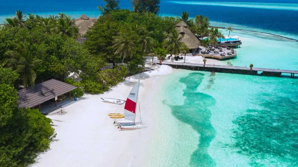 Komandoo Island Resort, activities, Lhaviyani Atoll, Maldives, Beach watersports area low-altitude aerial: red-sailed catamaran beached on the white sand fringe, the watersports thatched shelter at the left, the dive-centre thatched pavilion and lagoon swimming pool at the right edge, dense coconut canopy through the centre, the jetty extending to the right with darker reef patches showing through the turquoise lagoon.