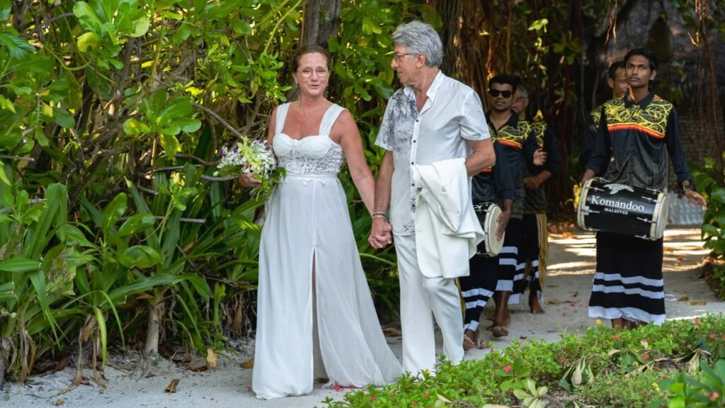 Komandoo Island Resort, lifestyle, Lhaviyani Atoll, Maldives, Maldivian wedding processional on the property: bride in white gown holding a bouquet walking arm-in-arm with the groom, three Maldivian musicians behind in traditional black-with-yellow-trim costume carrying boduberu drums (one drum visibly labelled with the property name), the tropical vegetation framing the sandy path, the wedding-and-celebration programme captured in property-signature staging.