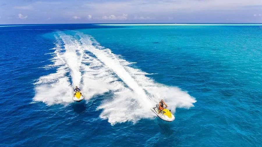 Kudadoo Maldives Private Island, activities, Lhaviyani Atoll, Maldives, Jet ski excursion aerial: two riders trailing wake patterns across deep cobalt ocean, the turquoise lagoon edge visible at the horizon, the non-motorised plus motorised watersports programme included within the all-inclusive plan arithmetic.
