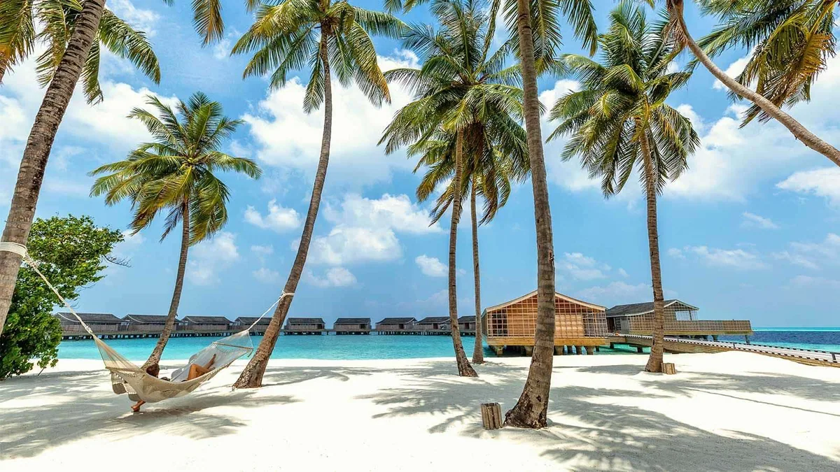 Kudadoo Maldives Private Island, lifestyle, Lhaviyani Atoll, Maldives, Beach hammock view: white sand foreground with a hammock strung between two tall coconut palms (single guest resting), the Ocean Residence row plus The Retreat building visible across the lagoon at the mid-distance, the curving overwater villa row extending across the lagoon at the back, the signature Kudadoo composition captured in single frame from the island side.