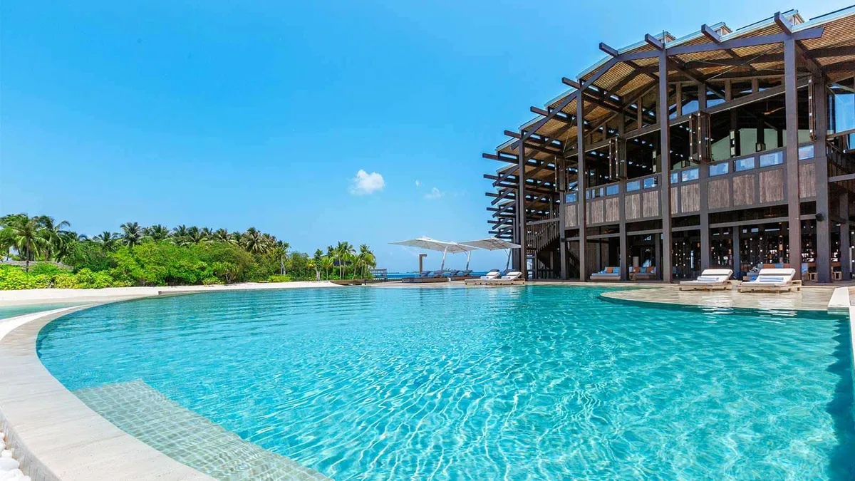 Kudadoo Maldives Private Island, architecture, Lhaviyani Atoll, Maldives, The Retreat building from the pool angle: two-storey timber-clad architectural pavilion with the distinctive louvered solar-panel roof and timber-pillared open-air structure, the chamfered-curve amoeba infinity pool extending into the foreground, sun loungers under parasols at the pool edge, white sand and dense palm canopy enclosing the scene.