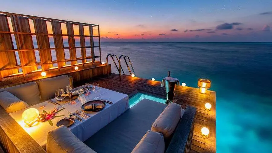 Kudadoo Maldives Private Island, dining, Lhaviyani Atoll, Maldives, Private overwater in-residence dining at twilight: table for two set with white-linen runner, a sphere lantern, flower garnish, champagne in an ice bucket, deep cushioned banquette seating wrapping the table, glowing lanterns along the perimeter of the deck, the plunge pool with underwater lighting beneath, pastel pink-violet-blue sunset sky over calm ocean.