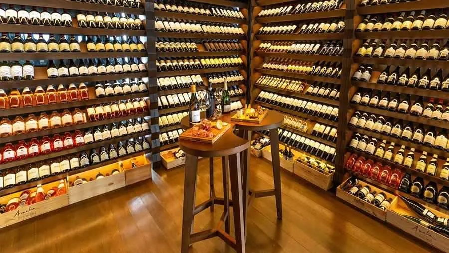 Kudadoo Maldives Private Island, dining, Lhaviyani Atoll, Maldives, Wine cellar tasting room: walls fully lined with hundreds of bottles in timber-rack arrangements, a central round high-table set with charcuterie boards plus cheese-and-fruit selection plus open bottles for tasting, warm timber floor, accent-lit shelves, the all-inclusive plan's premium-wine programme captured in property-signature staging.