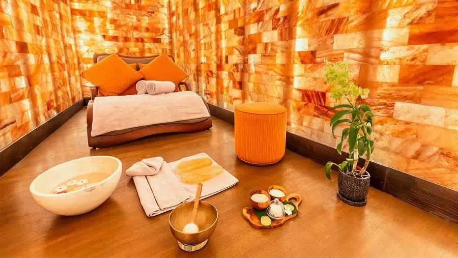 Kudadoo Maldives Private Island, spa, Lhaviyani Atoll, Maldives, Spa treatment room: backlit Himalayan salt-brick wall cladding in a golden-amber glow, a low timber-platform massage bed with orange and peach pillows plus folded towels, an orange velvet round stool, potted orchid plant in the corner, foreground ritual setup with copper-tone water bowl, folded towel, brass singing bowl, and small tasting tray of ramekins.