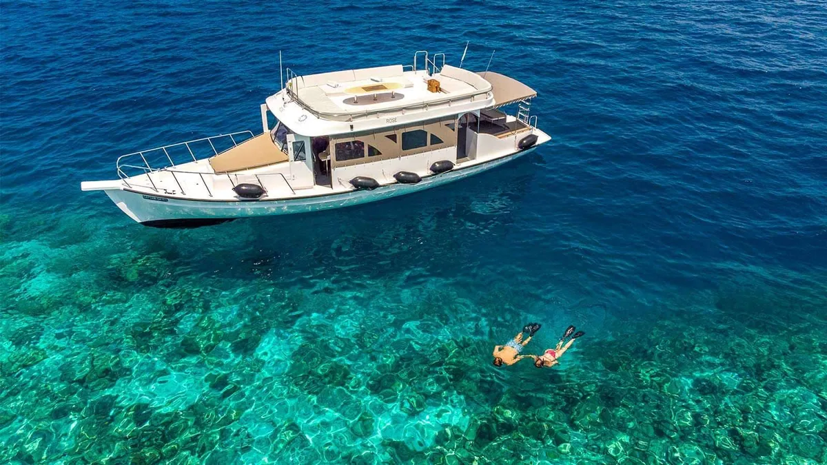 Kudadoo Maldives Private Island, activities, Lhaviyani Atoll, Maldives, Property dhoni snorkel excursion: top-down aerial of the Rose dhoni anchored over a coral-patch lagoon site, a snorkelling couple in the crystal water nearby, visible coral patterns beneath the surface, the included-everything dive-and-snorkel programme captured in property-named-vessel framing.