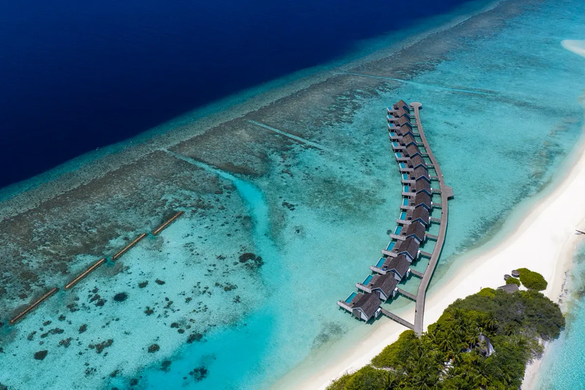 Niva Kuramathi Maldives (formerly Kuramathi Maldives), aerial, Alif Alif Atoll (North Ari), Maldives, Long arc water bungalow row drone aerial along the reef-edge boardwalk, 25 thatched bungalows on stilts following the lagoon-edge sweep, dense palm canopy on the 1.8-km Kuramathi island visible at right.