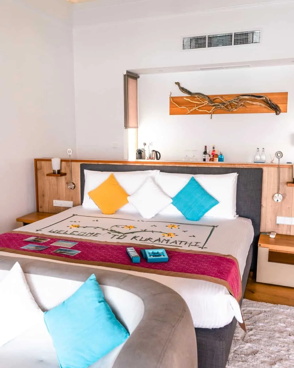 Niva Kuramathi Maldives (formerly Kuramathi Maldives), villa overwater, Alif Alif Atoll (North Ari), Maldives, Deluxe Water Villa bedroom interior, king bed dressed with a Welcome to Kuramathi embroidered runner and flower petals, turquoise and yellow accent throw pillows, sculptural driftwood-art panel above the headboard, wooden floors. Property-specific welcome detail.