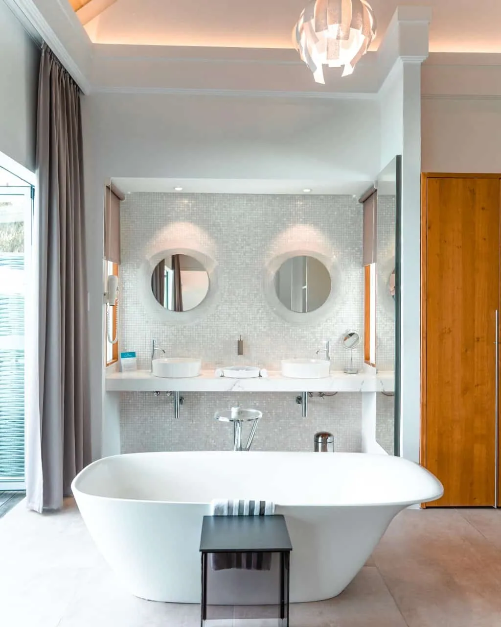 Niva Kuramathi Maldives (formerly Kuramathi Maldives), villa overwater, Alif Alif Atoll (North Ari), Maldives, Deluxe Water Villa bathroom, freestanding white tub centred under a sculptural pendant lampshade, twin round mirrors above the marble-shelf double sinks, white-mosaic backsplash, light-grey walls and timber accents.