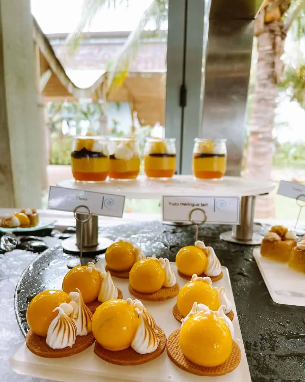 Niva Kuramathi Maldives (formerly Kuramathi Maldives), dining, Alif Alif Atoll (North Ari), Maldives, Farivalhu buffet dessert station, yuzu meringue tart row with golden mousse domes on round wafer bases, jars of compote on the tiered display behind, thatched-pavilion restaurant backdrop with palm fronds.