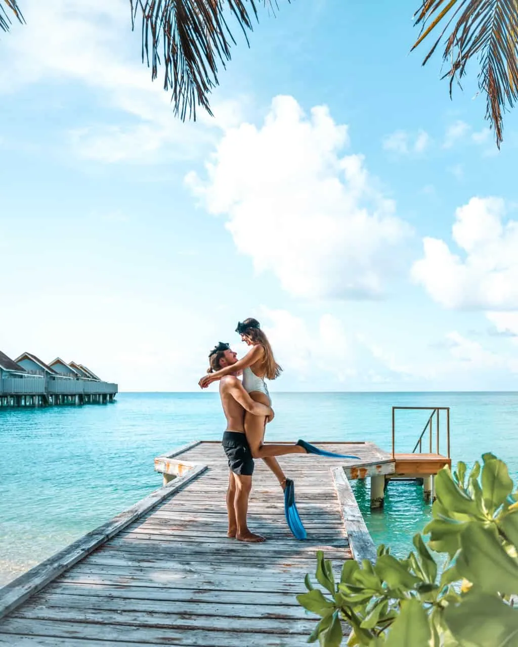 Niva Kuramathi Maldives (formerly Kuramathi Maldives), activities, Alif Alif Atoll (North Ari), Maldives, Snorkelling pair on a wooden jetty with blue fins, palm-frond canopy framing the foreground, the brown-roof overwater villa row reading along the left edge of the lagoon. The house-reef snorkel programme on the long-island perimeter.