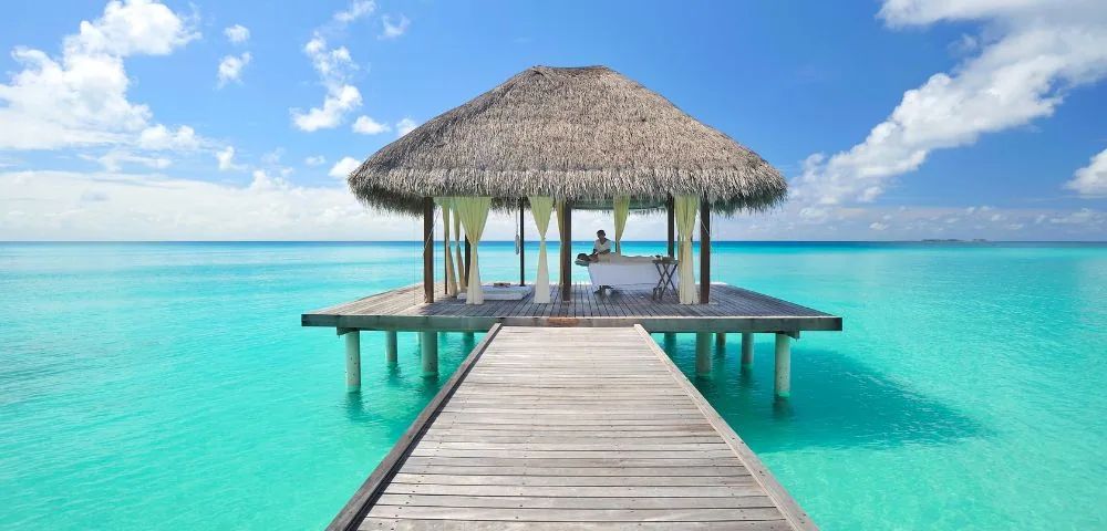 Niva Kuramathi Maldives (formerly Kuramathi Maldives), spa, Alif Alif Atoll (North Ari), Maldives, Overwater spa pavilion, thatched-roof pavilion at the end of a long wooden jetty extending into the turquoise lagoon, massage table inside with the therapist preparing the treatment. The property's premium-mid-tier wellness configuration in cinematic composition.