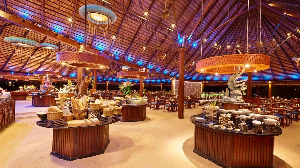 Kuredu Resort Maldives, dining, Lhaviyani Atoll, Maldives, Koamas buffet pavilion interior: cathedral-scale thatched roof with radial timber rafters illuminated by blue accent lighting along the joints, multiple round buffet stations with timber bases topped by suspended circular lampshade-feature lights, abundant warm-lit food displays, beach-style sand-and-cement floor, the mass-scale all-inclusive arithmetic captured in single property-signature frame.