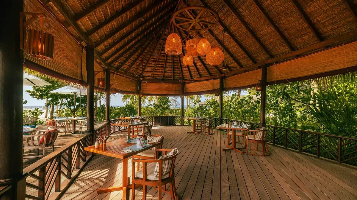 Kuredu Resort Maldives, dining, Lhaviyani Atoll, Maldives, Franco's Italian pavilion: open-air thatched-roof venue with vaulted timber ceiling, a signature cluster of woven-rattan basket pendant lights at the centre, wooden tables paired with orange-cushioned timber armchairs, the raised deck overlooking tropical palms with an ocean glimpse beyond, banister railing on the outer edge.