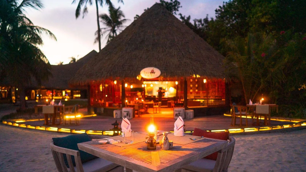 Kuredu Resort Maldives, dining, Lhaviyani Atoll, Maldives, Far East Asian-themed dining: conical thatched pavilion with red Chinese lanterns at the entrance, signage visible above, foreground table with twin folded napkins and an oil lantern set for service, perimeter LED strip lighting in warm yellow defining the sand-floor dining circle, dusky sky with palm silhouettes.