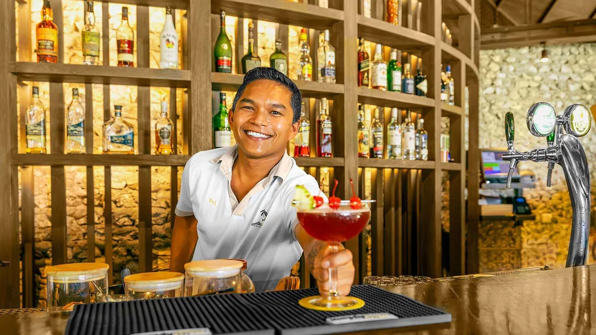 Kuredu Resort Maldives, bar, Lhaviyani Atoll, Maldives, Akiri Bar staffing: bartender in white polo carrying the Kuredu palm-tree logo presenting a margarita-glass cocktail garnished with cherries, behind him a curated coral-stone-clad backbar with timber alcoves displaying many liquor bottles, beer-tap apparatus at right, the multi-bar surface programme captured in property-branded framing.