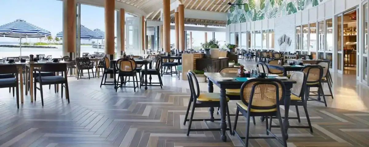 Le Méridien Maldives Resort & Spa, dining, Lhaviyani Atoll, Maldives, Turquoise interior: wide chevron herringbone parquet floor, multiple dark-wood dining tables paired with rattan-back chairs over yellow seat cushions, palm-leaf print mural panel on the back wall, the open glass-wall sides revealing the beach with the blue-and-white striped parasols visible outside, open kitchen visible at the far right.