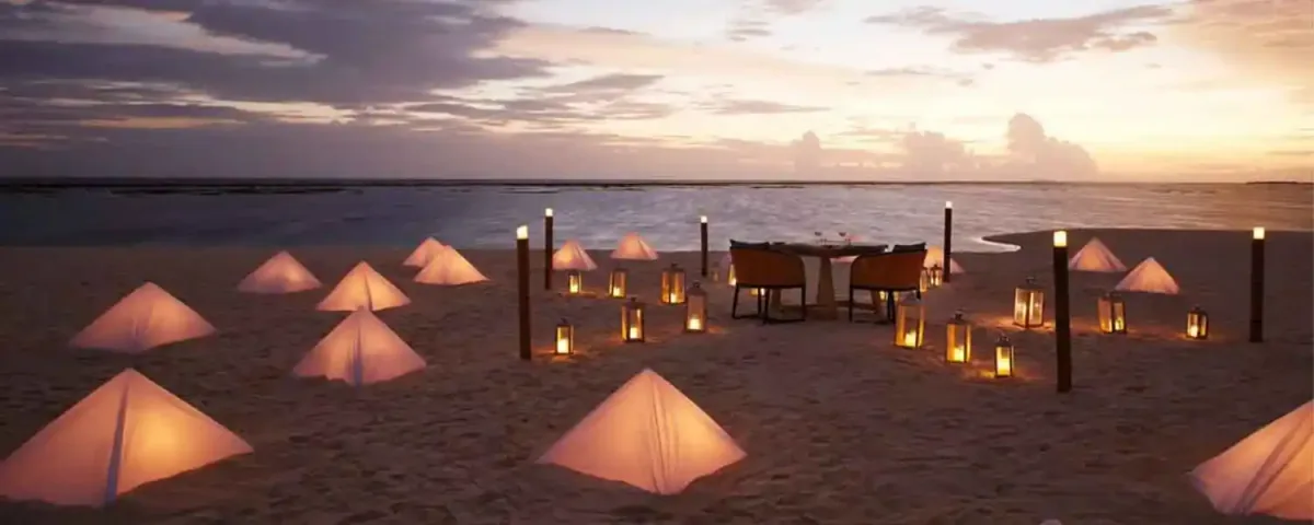 Le Méridien Maldives Resort & Spa, dining, Lhaviyani Atoll, Maldives, Private beach dining at dusk: a small dinner-for-two table on the white sand, surrounded by a constellation of glowing pyramid-tent paper lanterns plus tall floor lanterns in warm amber, the ocean and distant sandbar visible behind under a pastel dusk sky, the curated-private-experience programme captured in property-signature staging.