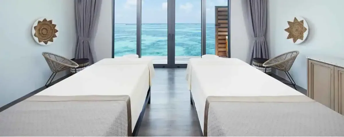 Le Méridien Maldives Resort & Spa, spa, Lhaviyani Atoll, Maldives, Explore Spa overwater couples treatment room: twin parallel massage beds with off-white linens facing a floor-to-ceiling picture window revealing the turquoise reef pattern below, two woven-rattan accent chairs on the sides, traditional Maldivian sun-disc wall plaques mounted on the side walls, the brand-signature Le Méridien wellness layer captured in the overwater-spa configuration.
