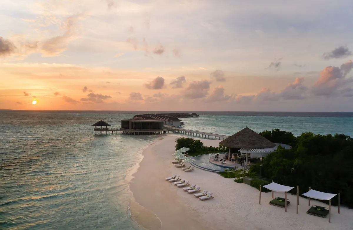 Le Méridien Maldives Resort & Spa, aerial, Lhaviyani Atoll, Maldives, Property aerial-oblique at sunset: the beach foreground with sun loungers and twin sand-cabana beds plus a thatched bar pavilion, an infinity pool at mid-frame, the over-water Velaa Bar & Grill structure visible on stilts (distinctive curved-overhang roof), distant over-water villa cluster on the horizon, golden-orange sunset sky over the lagoon.