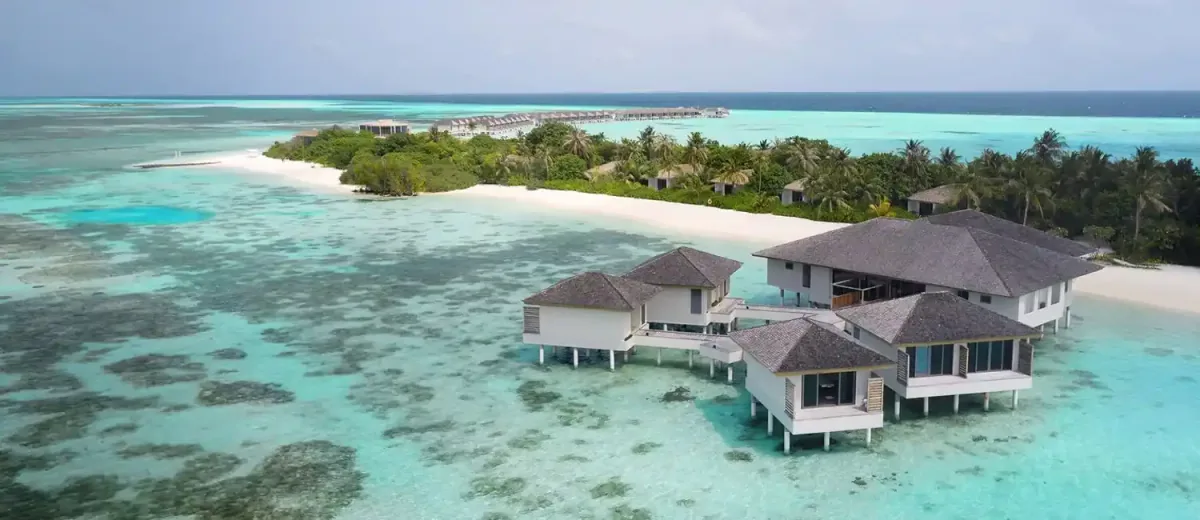 Le Méridien Maldives Resort & Spa, aerial, Lhaviyani Atoll, Maldives, Explore Spa overwater-pavilion cluster aerial: the four-pavilion shingled-roof spa cluster suspended over the reef-edge shallows on the north-east point of Thilamaafushi, the white sand-spit beach extending from the main island, the dense palm canopy of the main villa cluster, distant over-water villa row on the horizon edge.