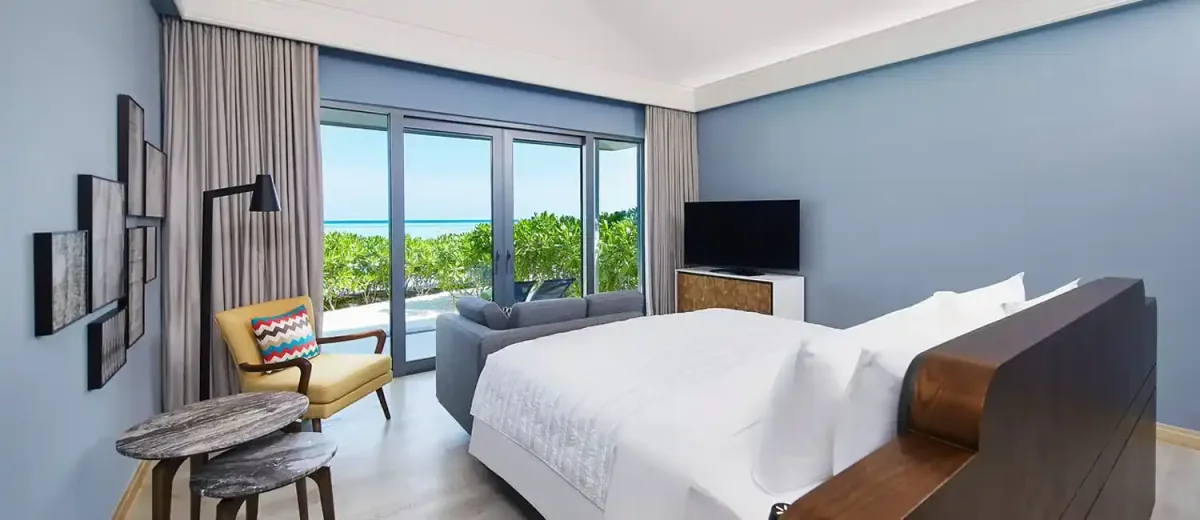 Le Méridien Maldives Resort & Spa, villa beach, Lhaviyani Atoll, Maldives, Beach Villa interior: Le Méridien dusty-blue painted walls (brand-signature palette), king bed with white linens against a walnut-veneer headboard, mustard-yellow Mid-Century-modern armchair with chevron-pattern cushion, multi-frame coral-rubbing art arrangement on the side wall, dark grey marble side-table cluster, sliding doors revealing tropical greenery and the turquoise lagoon beyond.