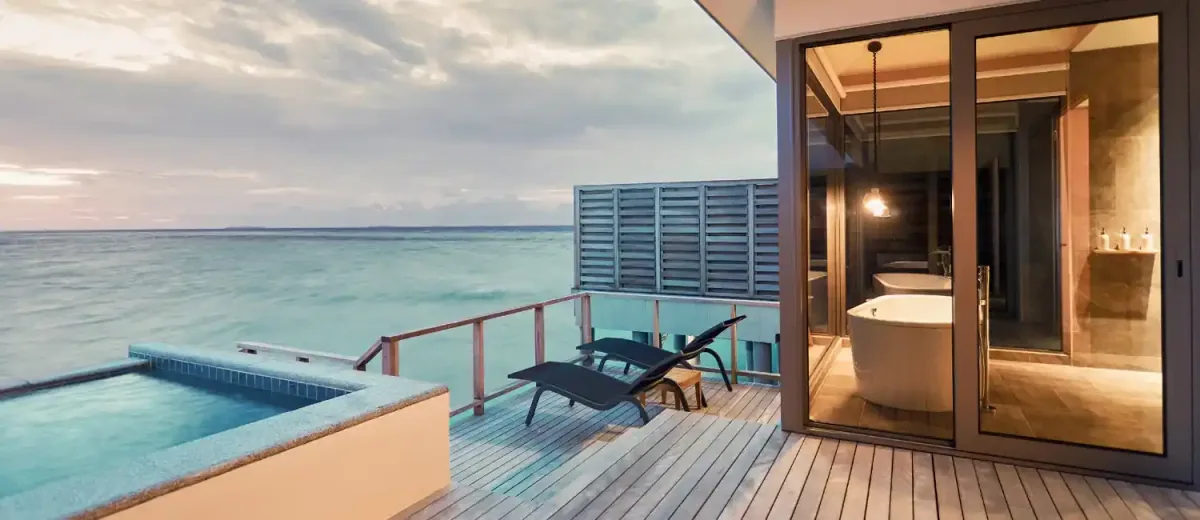 Le Méridien Maldives Resort & Spa, villa overwater, Lhaviyani Atoll, Maldives, Sunset Overwater Pool Villa private deck detail: square plunge pool with infinity edge cantilevered over the calm lagoon, single black chaise lounger on the timber deck, slatted timber privacy screen, the interior bathroom visible through sliding glass doors (freestanding tub, double vanity, pendant light), pastel-pink dusk sky.
