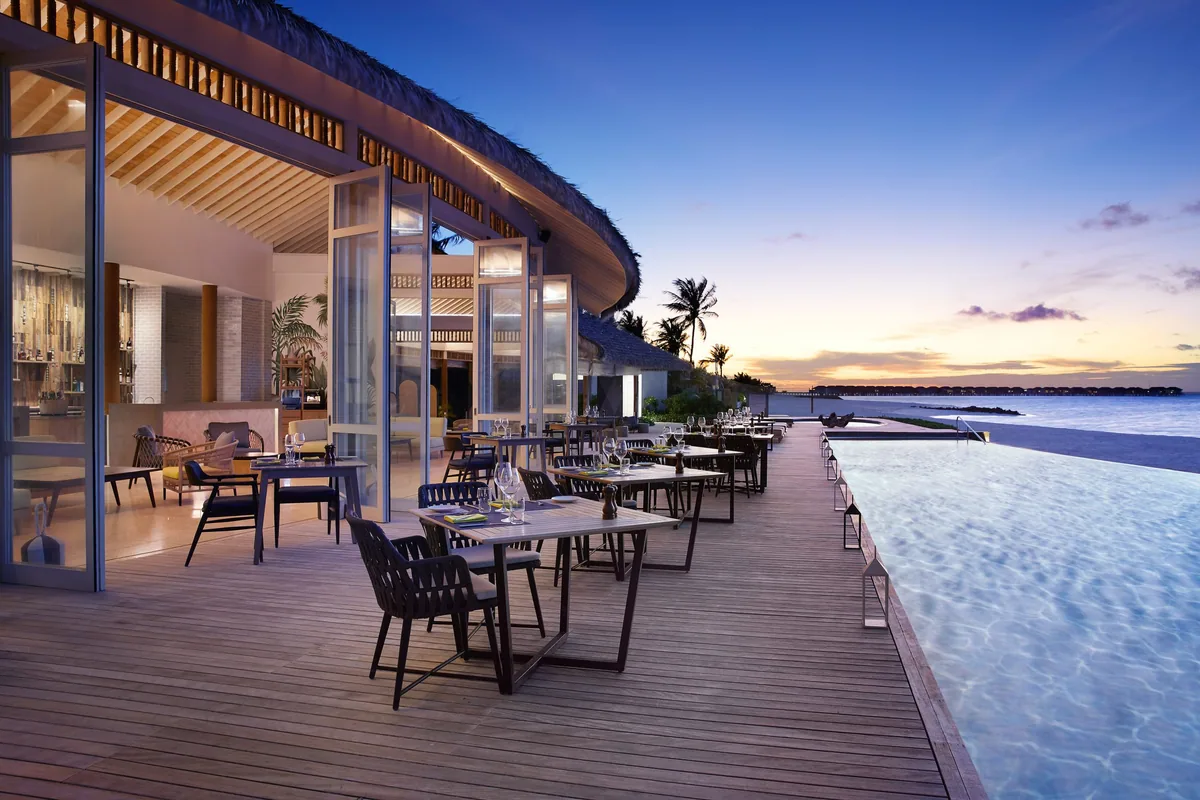 Le Méridien Maldives Resort & Spa, dining, Lhaviyani Atoll, Maldives, Velaa Bar & Grill at dusk: the signature curved overhanging thatched-roof pavilion on the pool-side terrace, timber-decked dining surface with tables and woven-rope dining chairs, the illuminated infinity pool extending to the right of the deck, distant over-water villa cluster on the horizon, soft peach-and-lavender dusk sky.