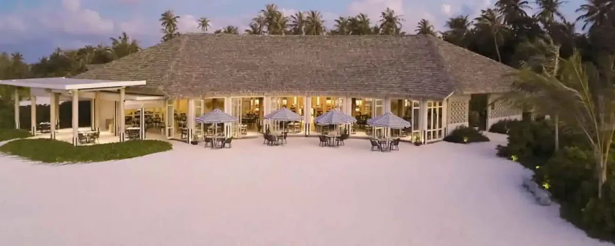 Le Méridien Maldives Resort & Spa, dining, Lhaviyani Atoll, Maldives, Turquoise pavilion beach-front exterior: low-pitched shingle-roof colonial-style pavilion with white columns and full-height glass walls revealing the warm-lit dining interior, four outdoor terrace tables under blue-and-white striped parasols on the white sand, the Le Méridien French-coastal Mediterranean motif (the striped parasols are the brand-signature beach-club element).