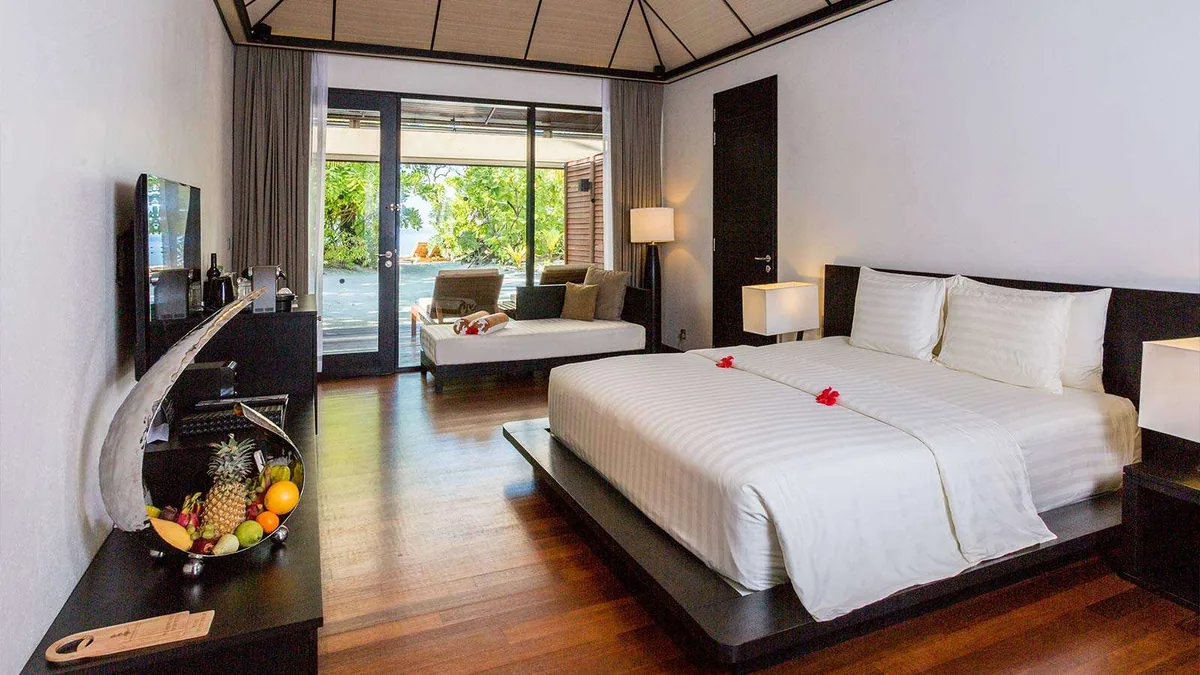 Lily Beach Resort & Spa, villa beach, Alif Dhaal (South Ari) Atoll, Maldives, Beach Villa interior with the dark-hardwood floor, king bed dressed in white linen with red frangipani trail, and the daybed alcove opening to the beach foliage.