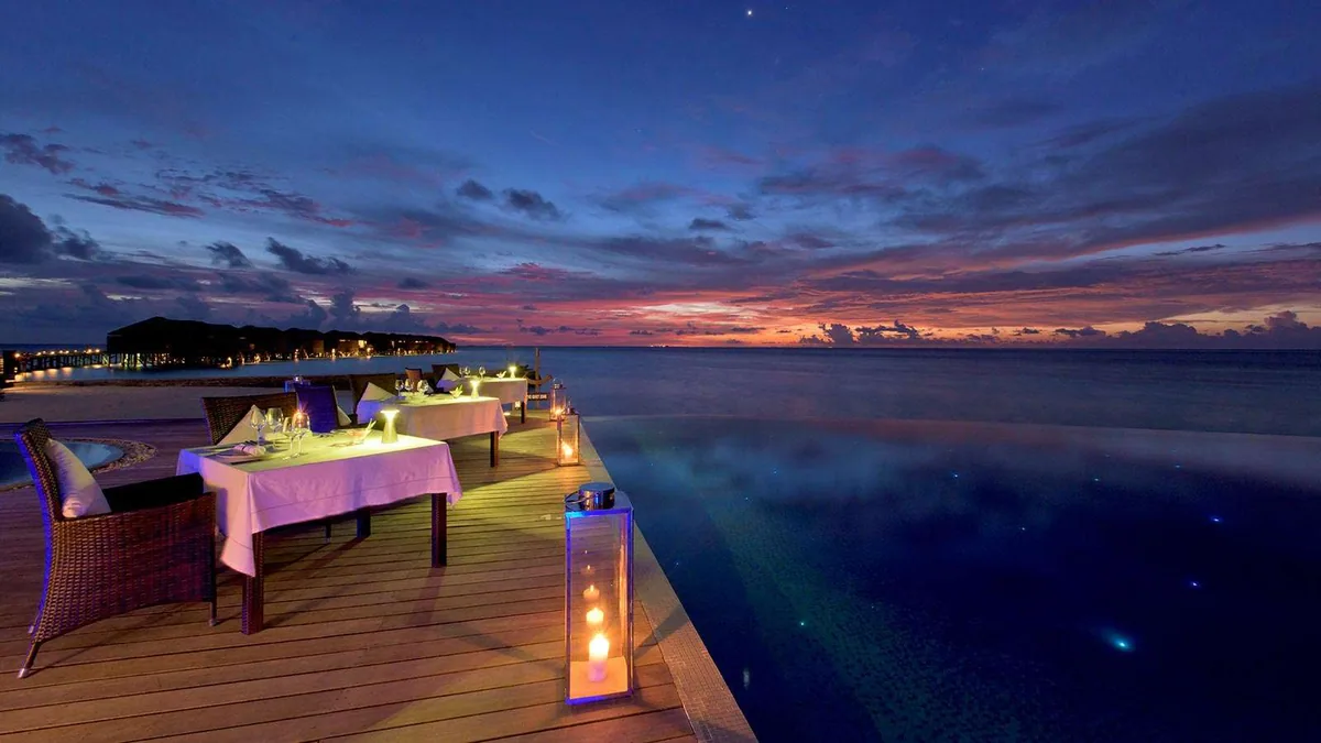 Lily Beach Resort & Spa, dining, Alif Dhaal (South Ari) Atoll, Maldives, Aqva pool-edge restaurant at twilight with the wicker armchair table line along the infinity pool and the candle lantern row at the deck edge.