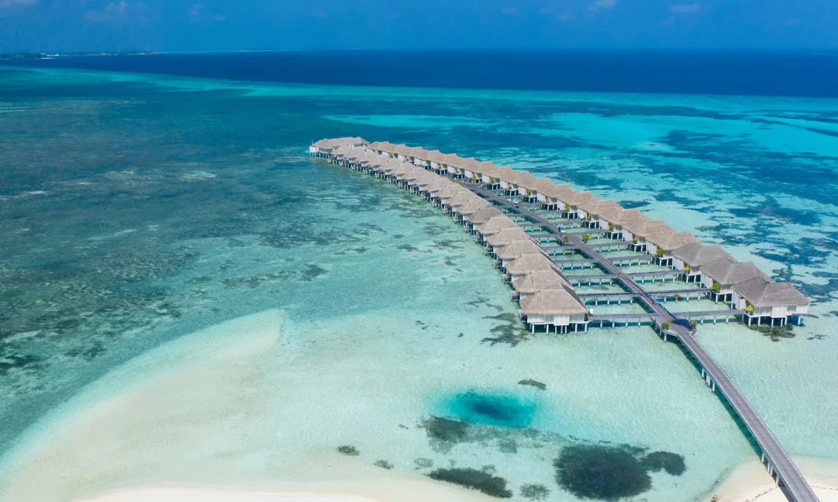 LUX* South Ari Atoll, aerial, Alif Dhaal (South Ari) Atoll, Maldives, Refurbished Water Villa row aerial with the parallel thatched-roof rank tracking along the long boardwalk and the sand-spit bottom-left with the deep-blue reef pothole.