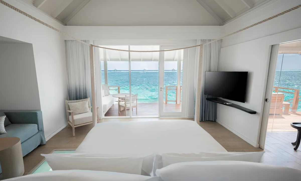 LUX* South Ari Atoll, villa overwater, Alif Dhaal (South Ari) Atoll, Maldives, Refurbished Water Villa bedroom with the vaulted white pavilion ceiling, rope-swag bunting, four-poster king bed, and the glass-floor panel revealing the lagoon at the bed line.