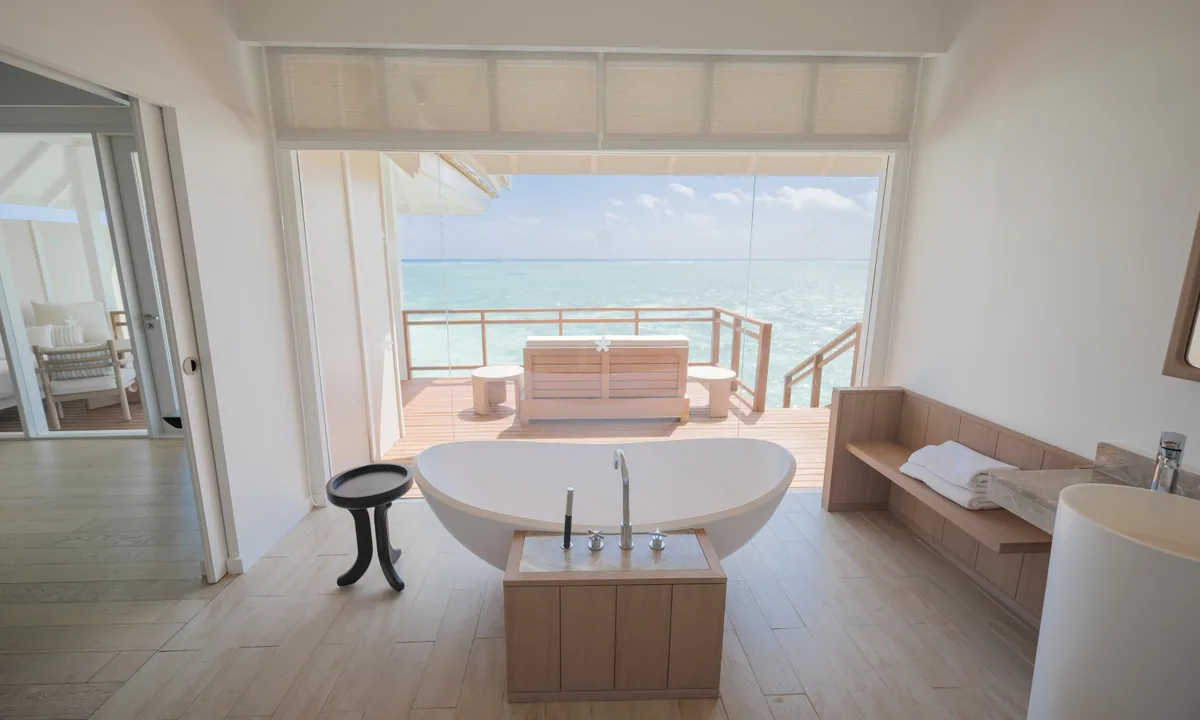 LUX* South Ari Atoll, villa overwater, Alif Dhaal (South Ari) Atoll, Maldives, Refurbished Water Villa bathroom with the asymmetric oval freestanding tub on the pale-pine plank floor and the deck-line opening to the L-shaped daybed and reef horizon.