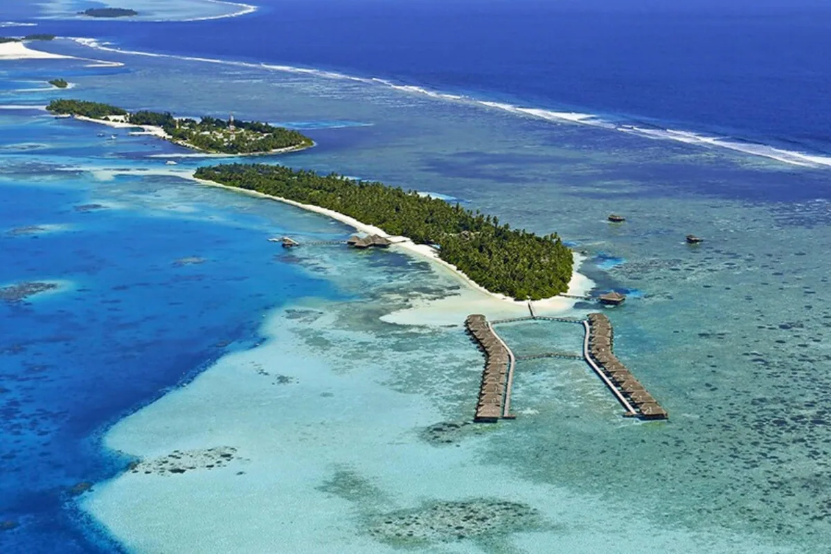 Medhufushi Island Resort, aerial, Meemu Atoll, Maldives, Medhufushi full island daylight aerial: the elongated palm-covered island stretching toward the horizon with the signature twin-arm overwater villa jetty extending into the lagoon (Y-shape configuration with the spa pavilion at the centre connecting two villa arms), turquoise reef formations visible beneath, smaller neighbour island in the far background.