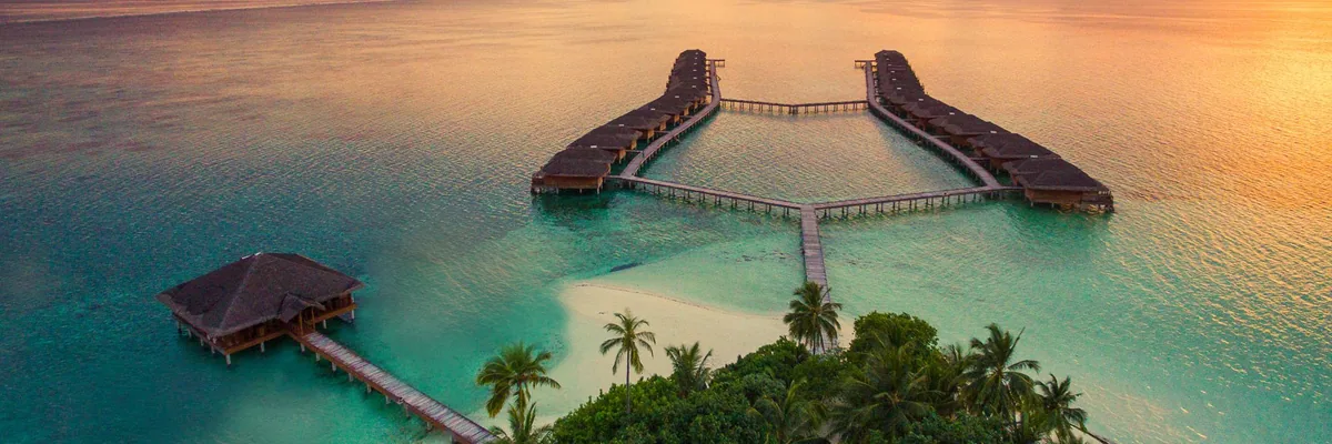 Medhufushi Island Resort, aerial, Meemu Atoll, Maldives, Medhufushi sunset aerial: same Y-shape overwater villa jetty (two parallel branches forming the top with a connecting walkway from the island) plus the separate overwater pavilion on the left edge, a small sand-spit beach extending from the island, golden-orange sunset reflection pooling across the lagoon, the property's signature composition captured in the golden hour.